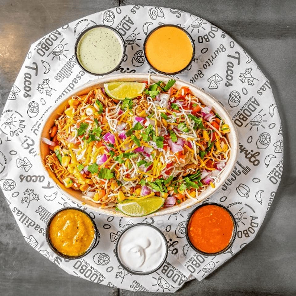 Overhead view of a colorful burrito bowl with lime wedges and five dipping sauces on a branded paper.